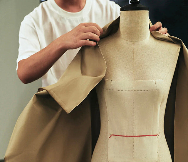 Person adjusting fabric on a mannequin wearing a beige garment.